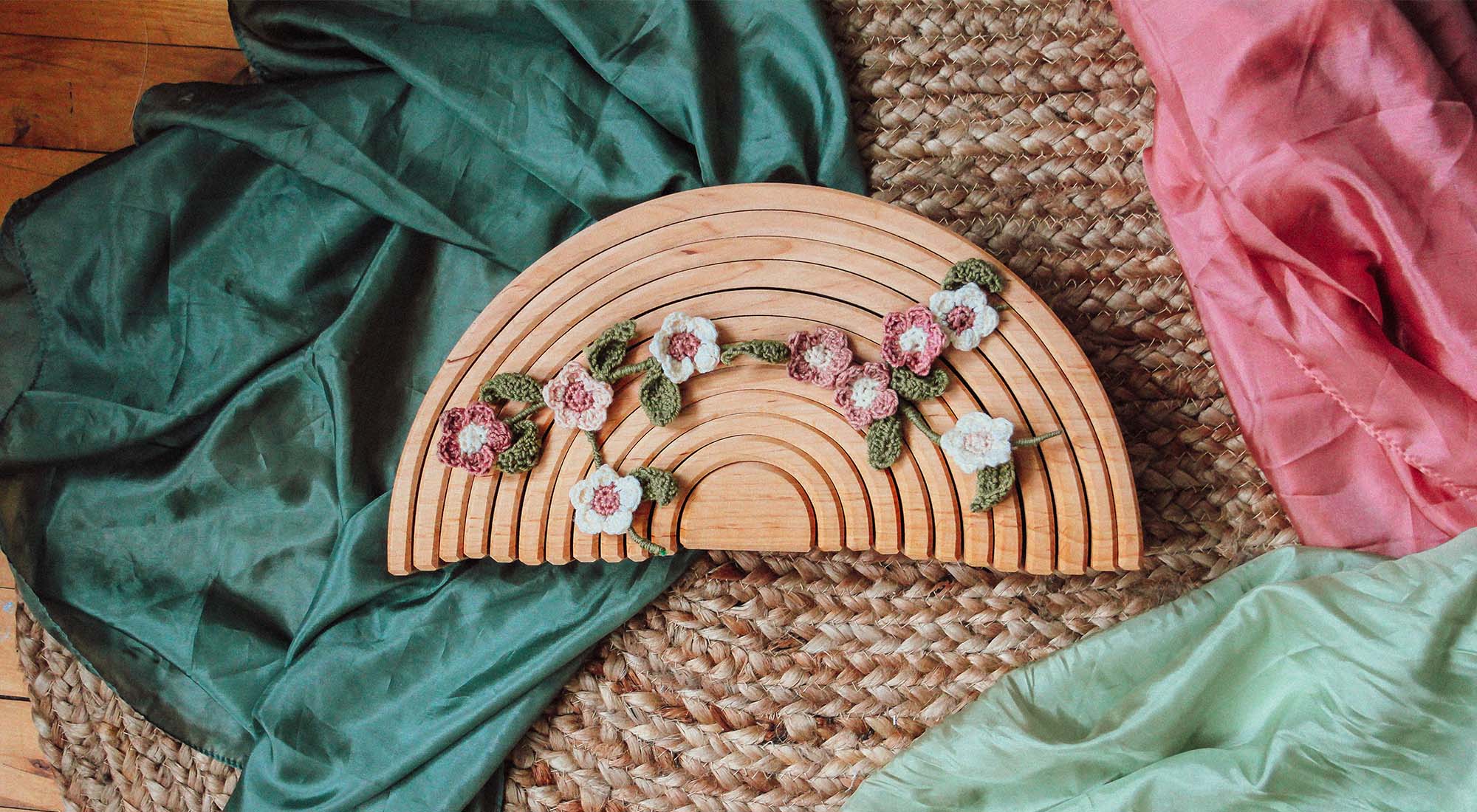 A crochet flower branch sits on a natural wooden rainbow with colorful playsilks.