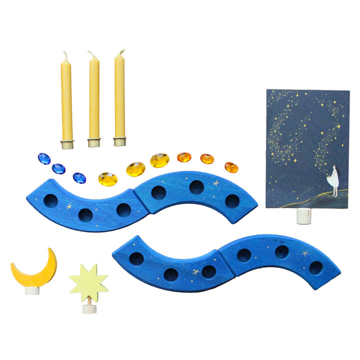 A Grimm's x Bella Luna Toys celebration ring set featuring a blue ring with hand drawn golden stars, 3 beesewax candles and brass candle holders, 1 moon decoration, 1 star decoration, 9 acrylic gems and a postcard holder with a celestial art card.