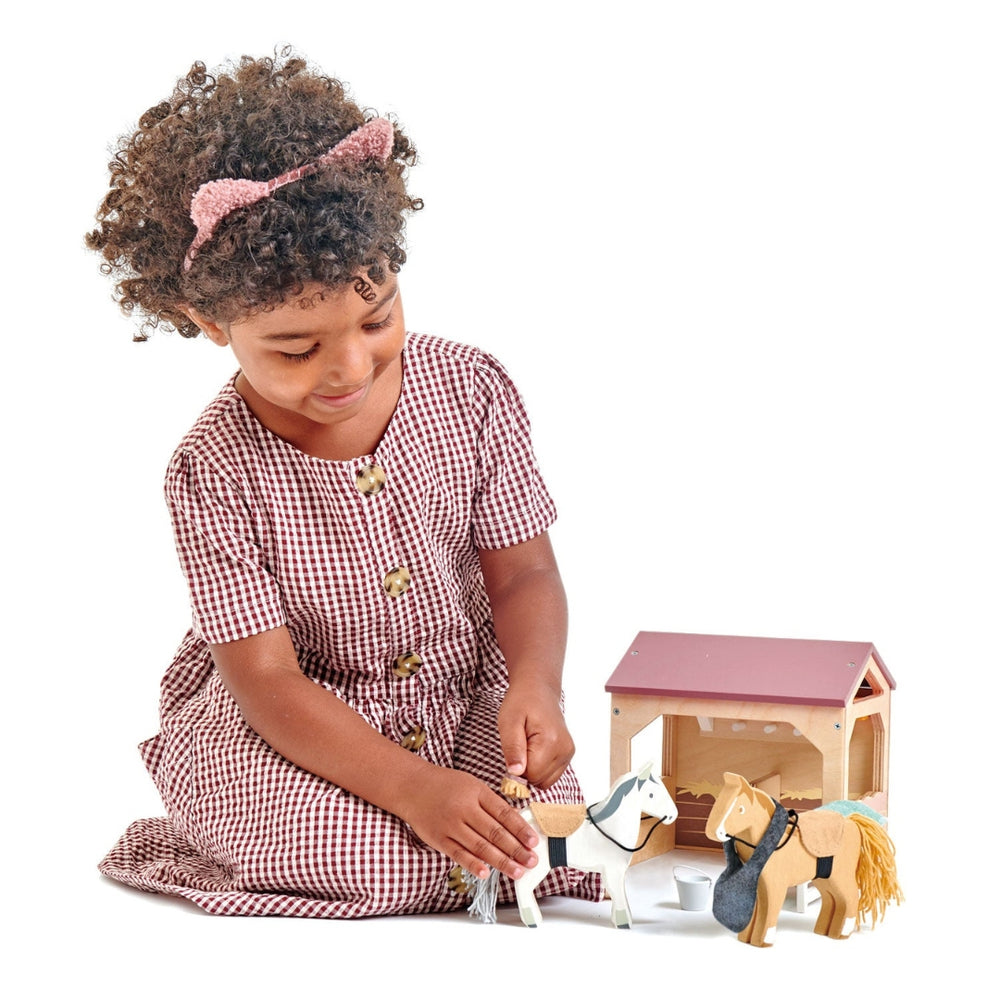 A young girl plays with a wooden horse and stable set by Tender Leaf Toys.