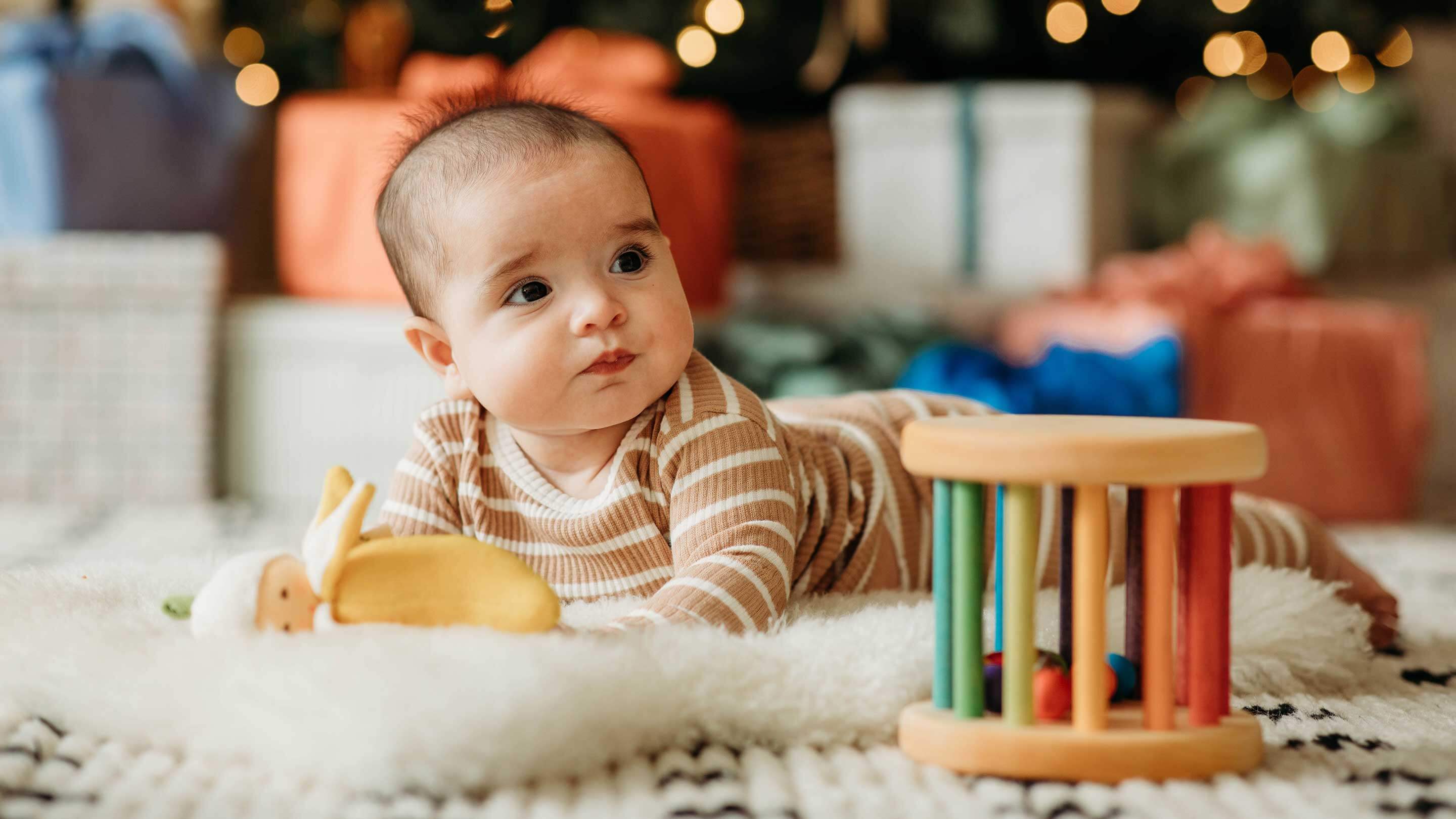 A baby laying on a sheepskin with a rainbow wooden toy and christmas presents in the background