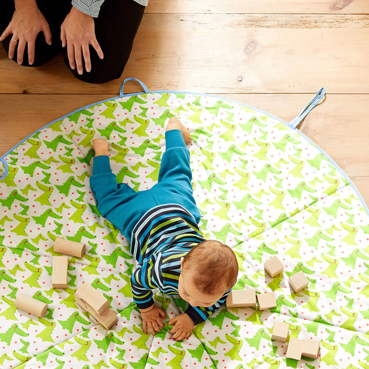 A baby crawls on a festive play mat surrounded by wooden blocks, with an adult's hands visible nearby.