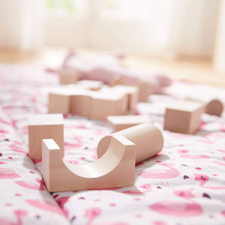 A soft pink blanket with playful patterns features scattered light wooden blocks of various shapes.