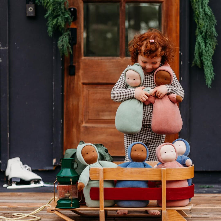 A girl holds two Bella Luna Toys Waldorf Dolls next to a child's sled, filled with more dolls.