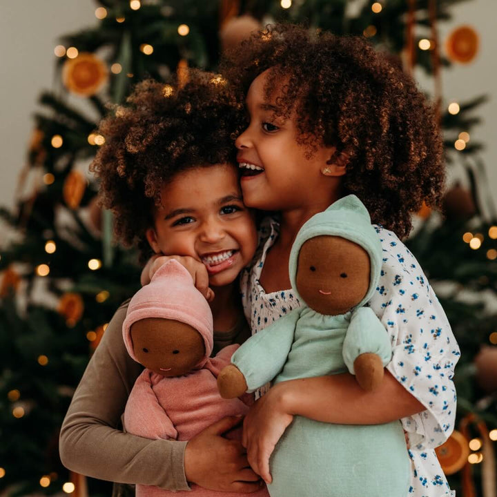 A brother and sister hug their Bella Luna Toys Waldorf Dolls in front of a Christmas tree.