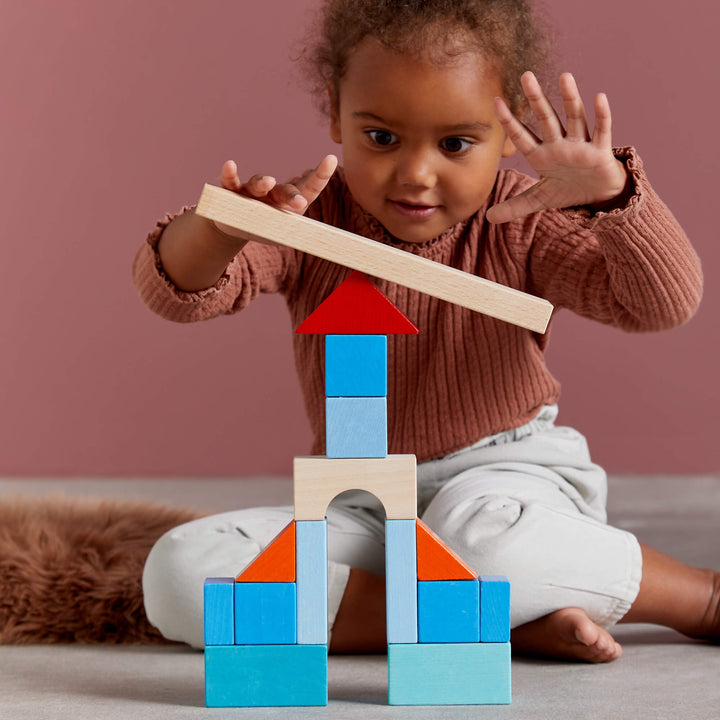 A child builds a colorful block tower on the floor, focusing intently as they balance a wooden piece on top.