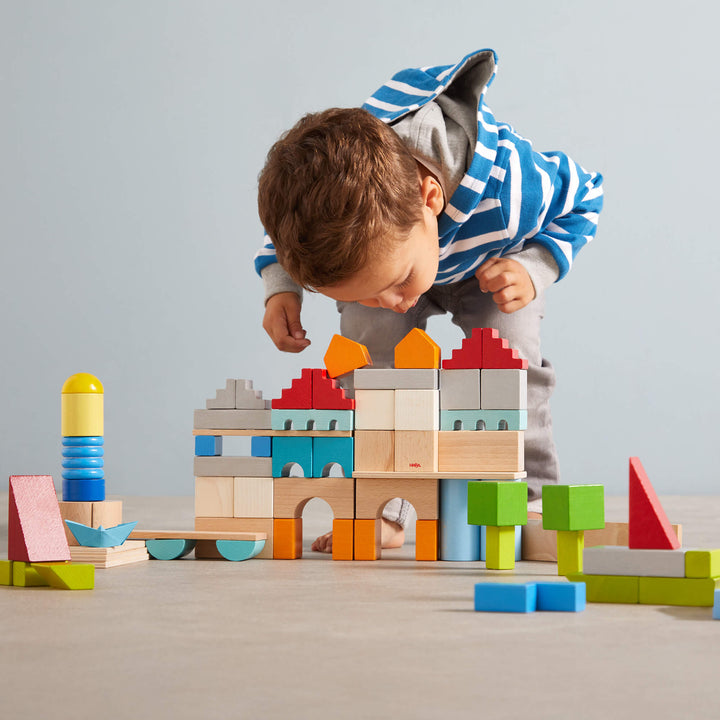 A young boy in a striped hoodie carefully inspects a colorful block castle he built on a light-colored floor.