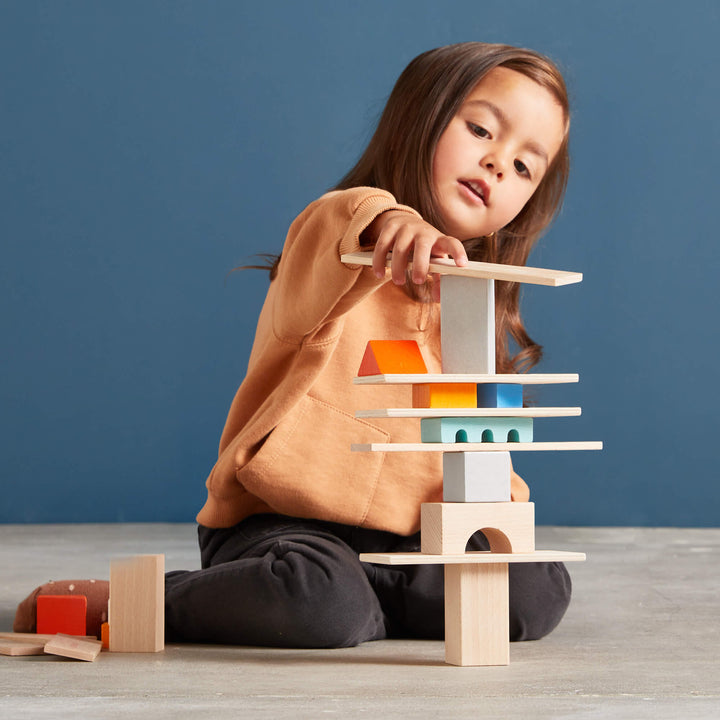 A young child focuses intently while stacking colorful wooden blocks into a tower on a light gray floor against a blue wall.