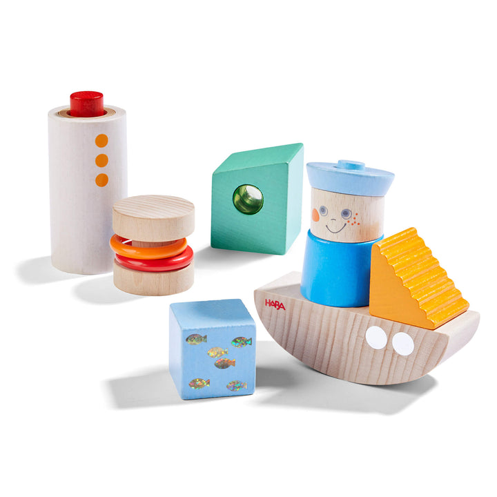 Discovery Building Blocks Boat Set with a tall natural cylinder with a red button and 3 orange dots, a short cylinder with 2 plastic orange and red rings, a teal rectangular shape with green plastic peep hole, a blue square with shiny fish, a half moon shape with 2 white dots, an orange triangle, and a blue wooden sailor