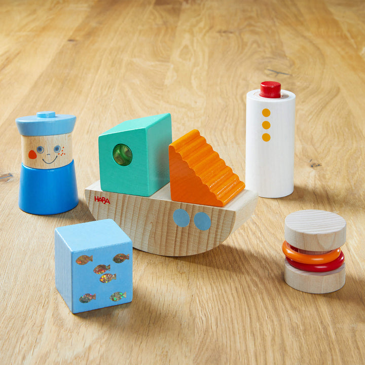 Discovery Building Blocks Boat Set with a tall natural cylinder with a red button and 3 orange dots, a short cylinder with 2 plastic orange and red rings, a teal rectangular shape with green plastic peep hole, a blue square with shiny fish, a half moon shape with 2 white dots, an orange triangle, and a blue wooden sailor on a wooden background