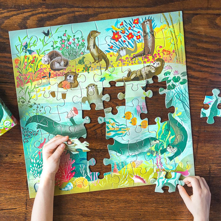 Childs hands placing pieces of Otters at Play 64 Piece Jigsaw Puzzle on wooden table