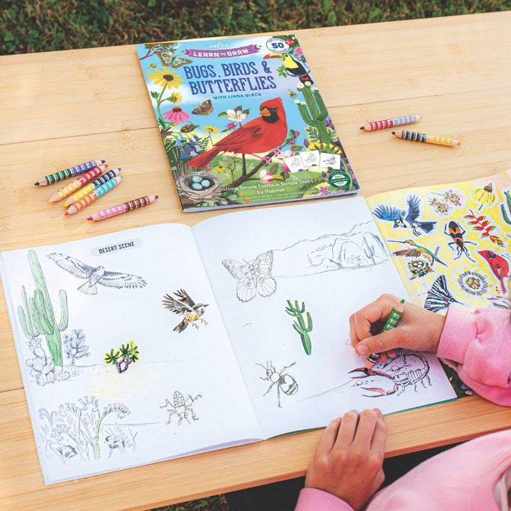 Child working in a wooden table outside with double-sided colored pencils coloring  in eeBoo’s Learn to Draw Birds and Butterflies book, featuring colorful illustrations, step-by-step drawing guides, and nature-themed designs for kids.