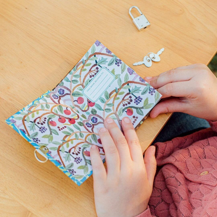 A close up of a child's hands holding the pocket-sized eeBoo Very Small Fairy Journal with whimsical fairy artwork by Gemma Koomen, gold foil accents, a metal lock, and two keys for secret writing open to he first page that shows a place to inscribe the book with your name. 
