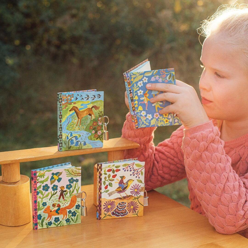 A younf child in a peach sweater looking at one of the journals with the three other pocket-sized eeBoo Very Small Fairy Journals with whimsical fairy artwork by Gemma Koomen standing upright on a wooden table.