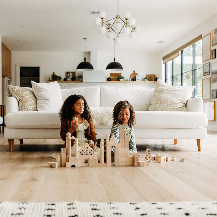 Two young girls sit on a wooden floor, smiling as they play with the Basic Building Blocks 102 Piece Extra Large Wooden Starter Set  in a bright modern living room. A sofa is in the background.