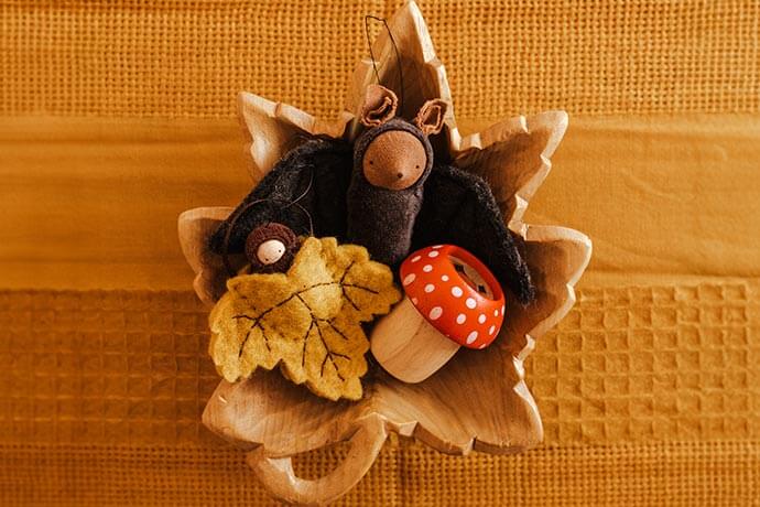 Wooden toy mushroom with felted leaf and felted toy bat in a wooden leaf bowl on a mustard colored tablecloth