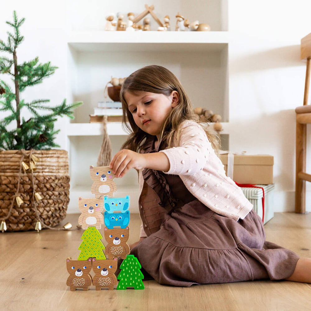 A young girl sits on the floor, carefully stacking colorful wooden bears and trees, with a festive background.