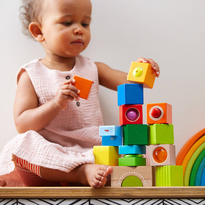 A toddler in a pink dress carefully stacks colorful wooden blocks, focused on building a playful tower.