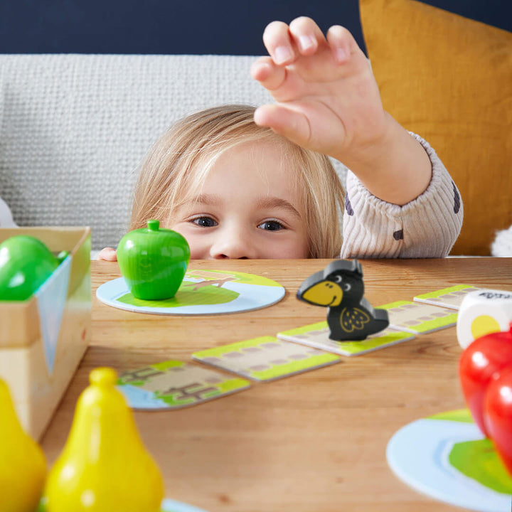 A young child with light hair reaches for a green apple during a colorful board game spread on a wooden table.