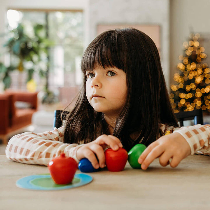 A young girl with long dark hair sits at a table, holding colorful toy fruits from the game First Orchard, with a Christmas tree softly glowing in the background.