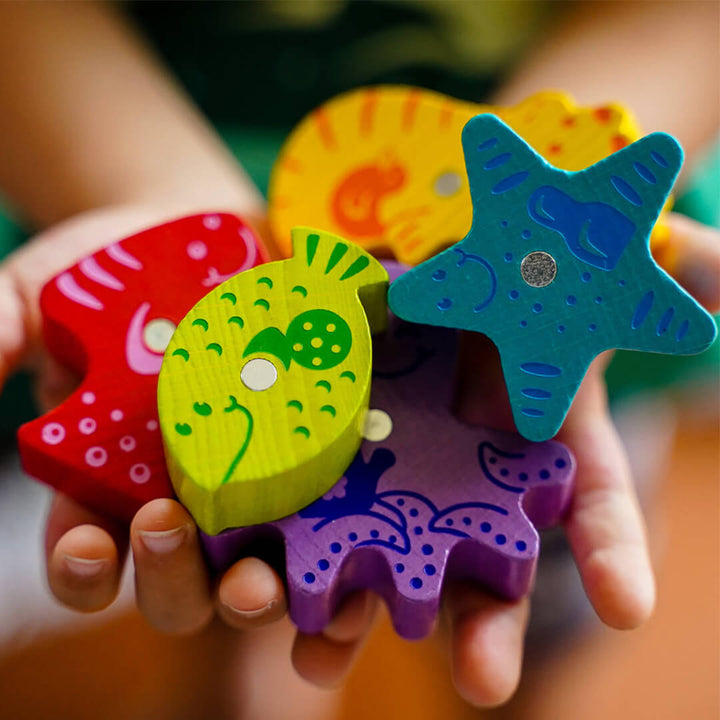 A child's hands holding a bright set of colorful wooden game pieces from Here, Fishy, Fishy! game including fish and starfish