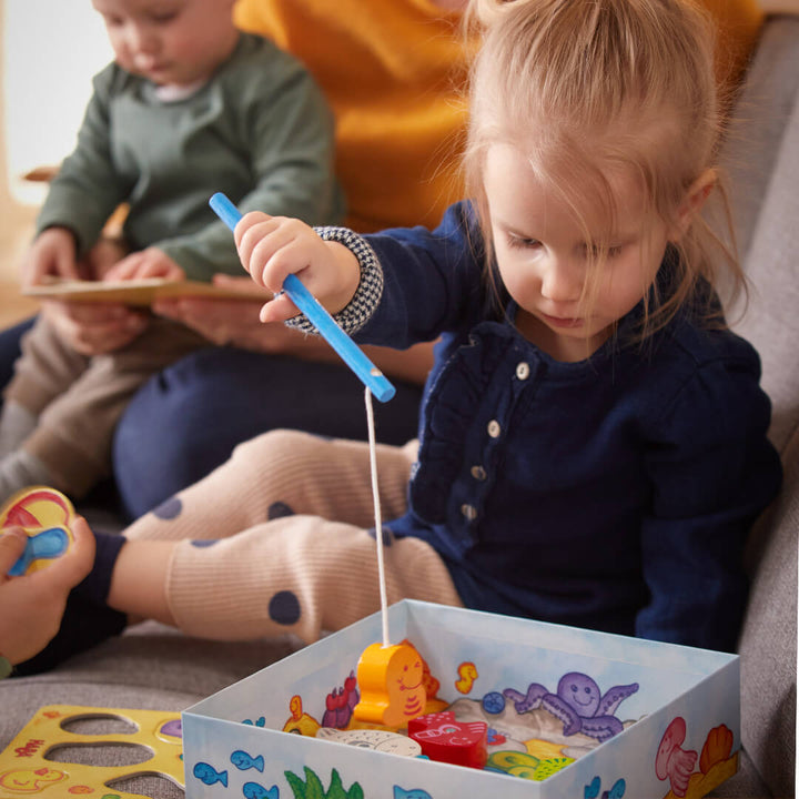 A young girl in a blue sweater plays with Here, Fishy, Fishy! Magnetic Game, while a baby and woman engage nearby on a sofa.