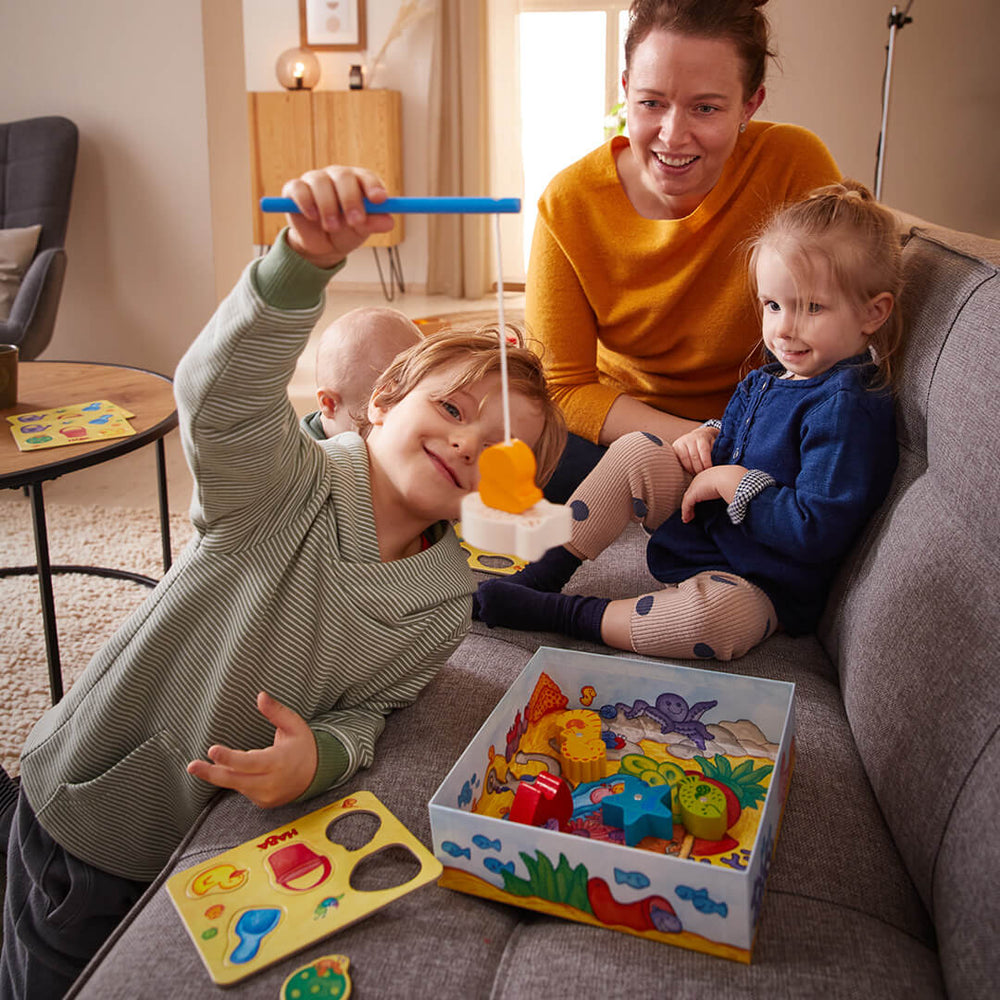 A woman and two children on a couch while the boy holds a magnetic fishing pole with a wooden fish attached 