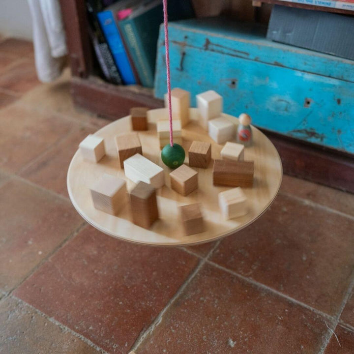 The Grapat Wooden Balance Board – filled with wooden blocks and the wooden green ball and a wooden figure hanging above a tile floor. A handcrafted toy for exploring balance, motion, and physics through open-ended play.