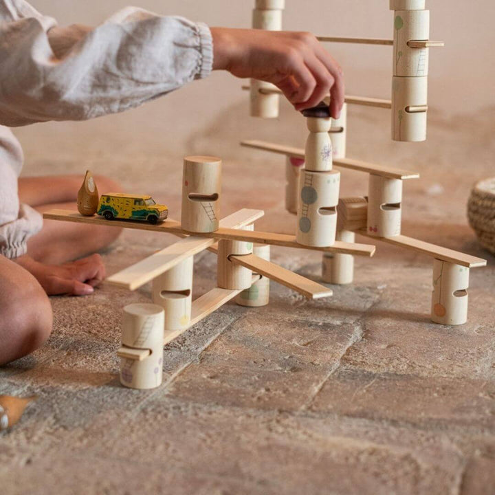 A child sets up the Grapat Woodland Stacking Tower Set with hand-painted wooden tubes and walkways, and has a toy car and the planks in a new design for open-ended building and imaginative play.