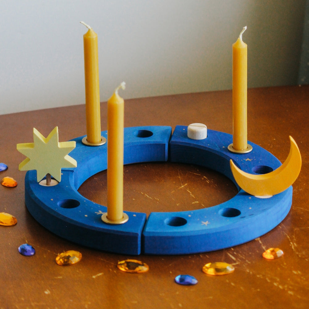 A Grimm's x Bella Luna Toys celebration ring set featuring a blue ring with hand drawn golden stars, 3 beesewax candles and brass candle holders, 1 moon decoration, 1 star decoration,  and 9 acrylic gems.