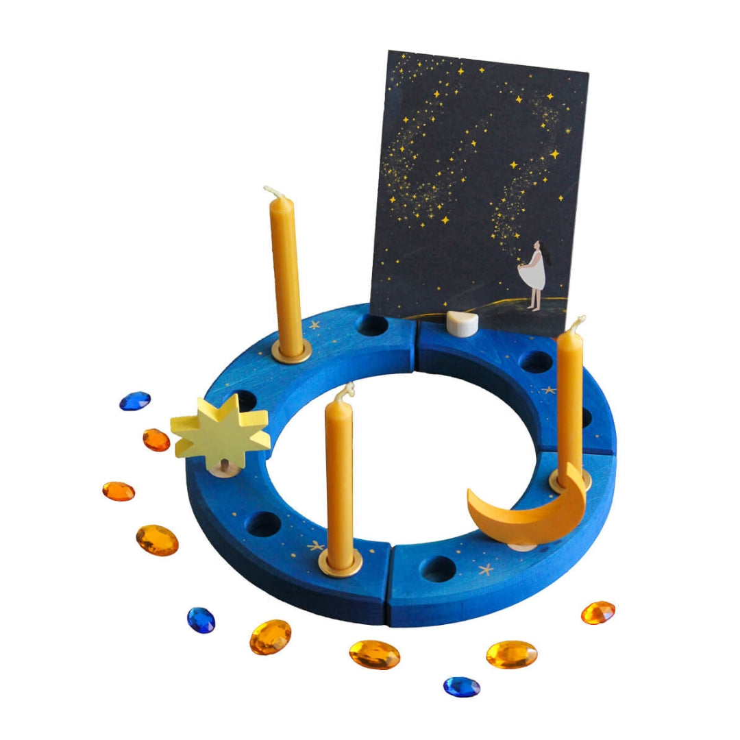 A Grimm's x Bella Luna Toys celebration ring set featuring a blue ring with hand drawn golden stars, 3 beesewax candles and brass candle holders, 1 moon decoration, 1 star decoration, 9 acrylic gems and a postcard holder with a celestial art card.