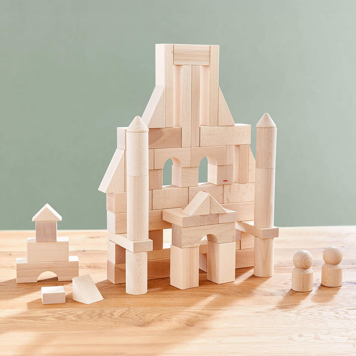 Basic Building Blocks 60 Piece Large Starter Set creating a wooden block castle featuring towers and arches, with smaller blocks and two round figures nearby on a wooden table.