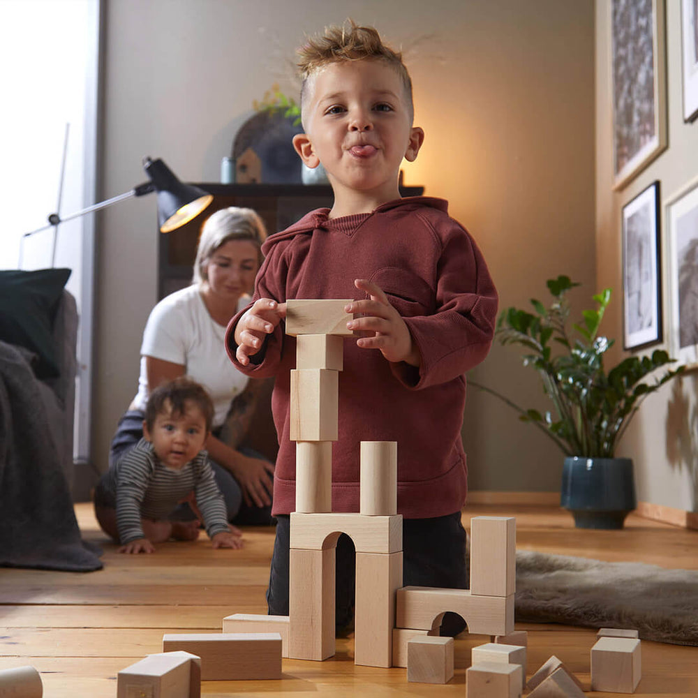 A playful young boy sticks out his tongue while balancing wooden blocks. A woman looks on as a baby crawls nearby.