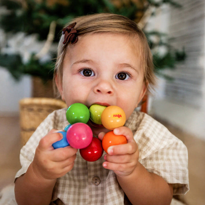 A toddler looking at the camera in a basket holds and chews on a Magica Wooden Clutching Toy made of colorful wooden balls.