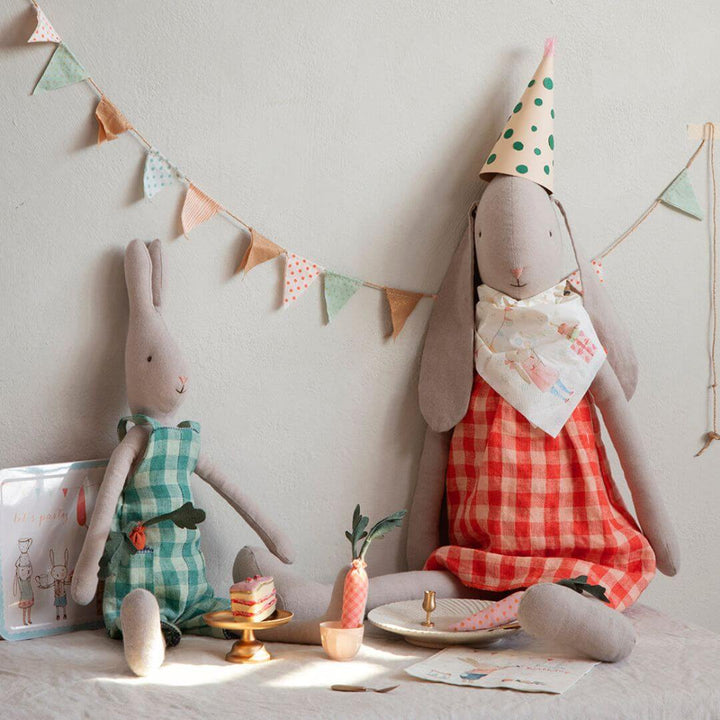 "A whimsical birthday party scene featuring two Maileg bunnies dressed in gingham outfits. One bunny, wearing an orange dress and a party hat, sits with a festive bib, while the other in a green dress joins the celebration. A miniature table is set with tiny cakes, a carrot, and decorations, with pastel bunting hanging in the background.