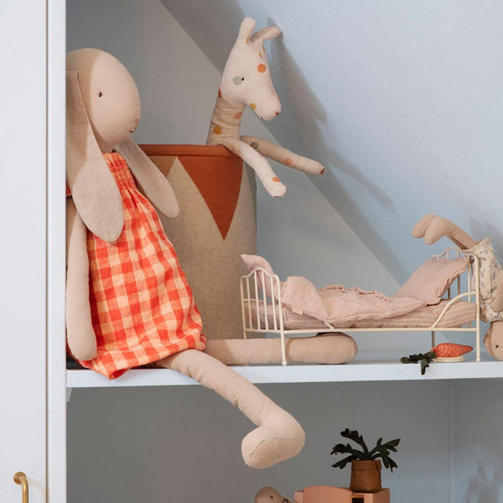 A charming nursery shelf display featuring a Maileg Bunny in an orange gingham dress sitting on the edge. A plush giraffe peeks out from a storage bin, while a tiny Maileg bunny rests in a miniature bed with a cozy blanket. A small potted plant adds a decorative touch to the whimsical scene.