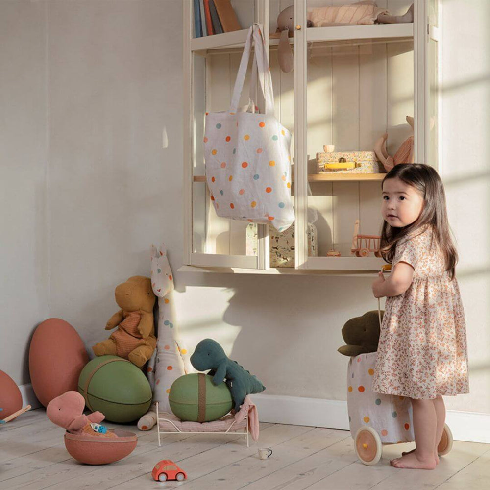 A playful scene featuring Maileg Gantosaurus plush toys, including a small Dark Petrol dino nestled inside an open egg, surrounded by other dinos in a cozy, vintage-inspired setting with a young girl holding a rolling polka dot bag.