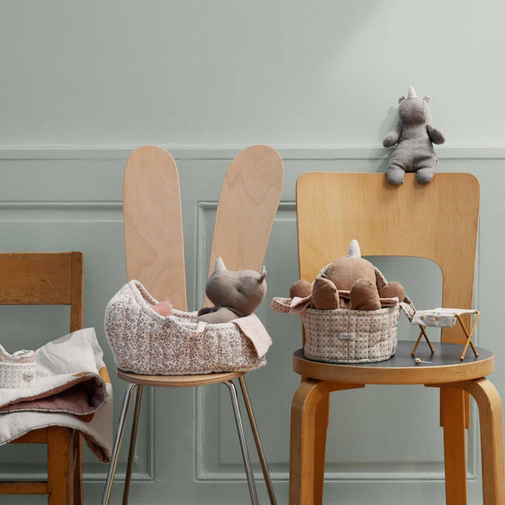 A whimsical nursery scene featuring two Maileg carry cots placed on wooden chairs, each holding a plush rhino tucked under a cozy blanket. Another small plush rhino sits on the chair back, adding to the charming and playful setting.