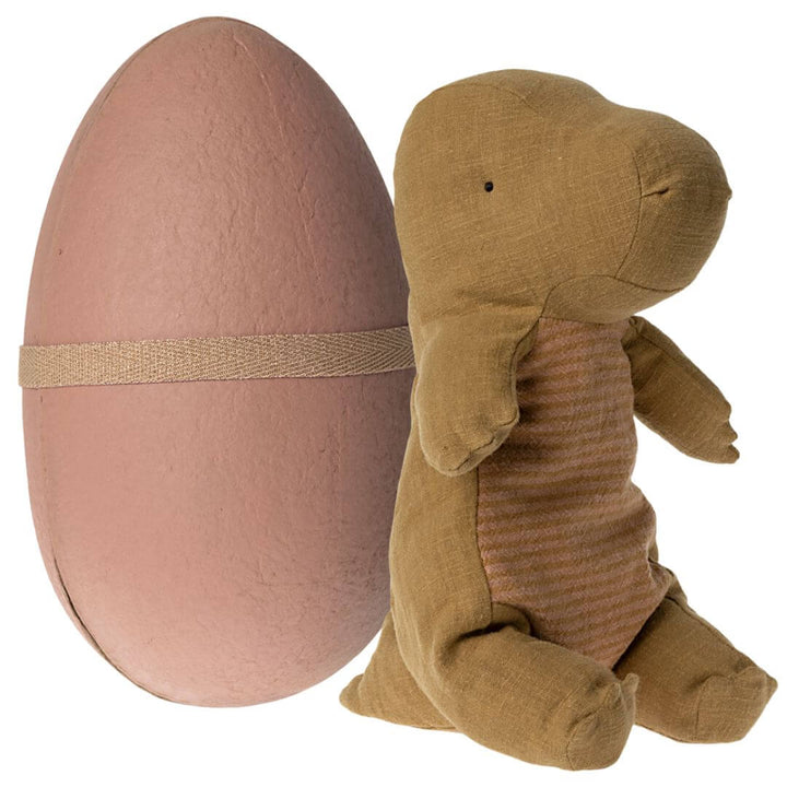 Maileg Medium Gantosaurus in Egg – Dark Ocher. A soft, plush golden-brown dinosaur with a striped belly, sitting beside its matching ocher egg-shaped shell secured with an elastic band.