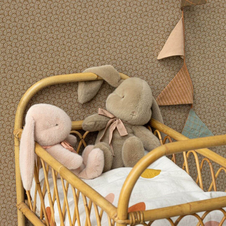 Maileg Medium Plush Bunnies in Dusty Brown and Powder snuggled in a miniature rattan crib with soft bedding and a garland hanging behind them on the wall. A cozy nursery scene perfect for Easter or gifting