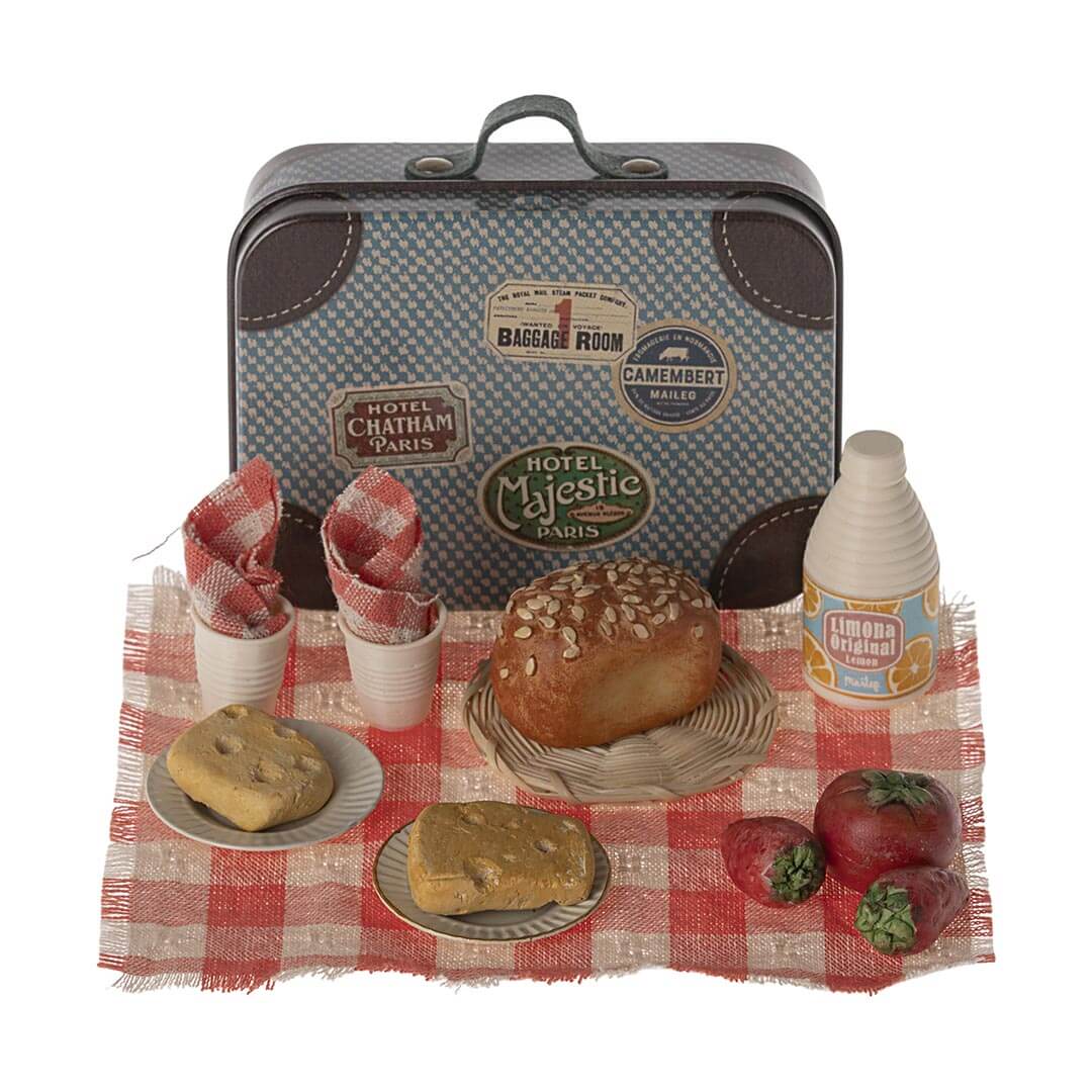 Maileg Mouse Sized Picnic Set with red and white checked blanket, a loaf of bread, 2 cups, 2 napkins, 2 stawberries, 1 tomato, 1 orange juice, 2 plates, 2 pieces of bread with wholes, and a woven charger with a blue patterned suitcase