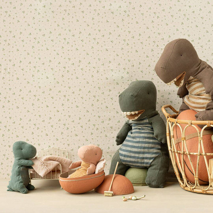 A cozy scene featuring Maileg Gantosaurus plush toys in various sizes, including a small rose-colored dino resting inside an open egg. Other dinos, dressed in striped outfits, are gathered around, with one in a basket. The setting has a charming, vintage-inspired background with floral wallpaper and miniature accessories.