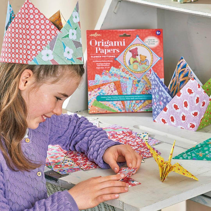 A young girl with an origami crown on her head folding origami paper next to an origami paper crane