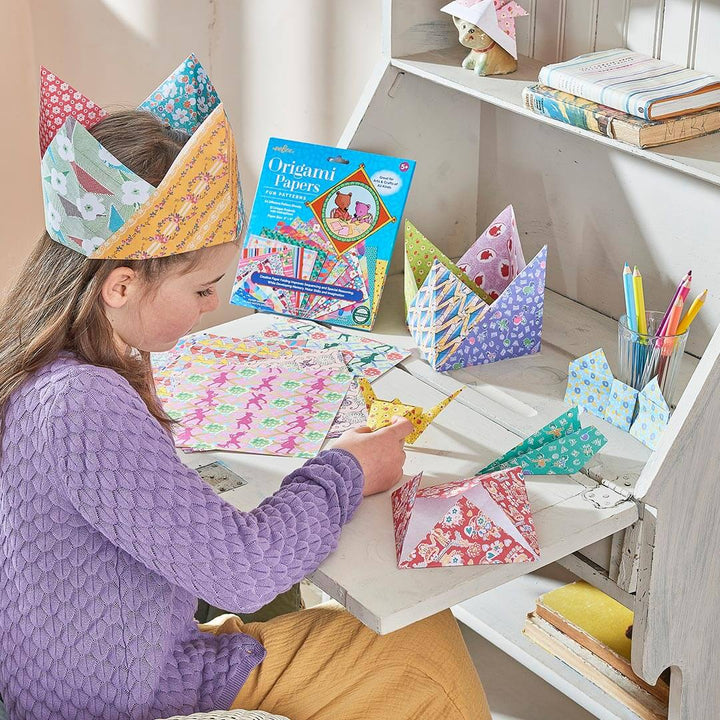 A young girl with origami paper crown folding an origami paper crane on a wooden desk