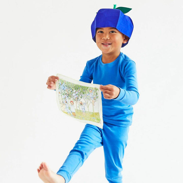 A child in blue pajamas and a blue silk blueberry hat is holding out a mini playsilk with a picture printed on it.