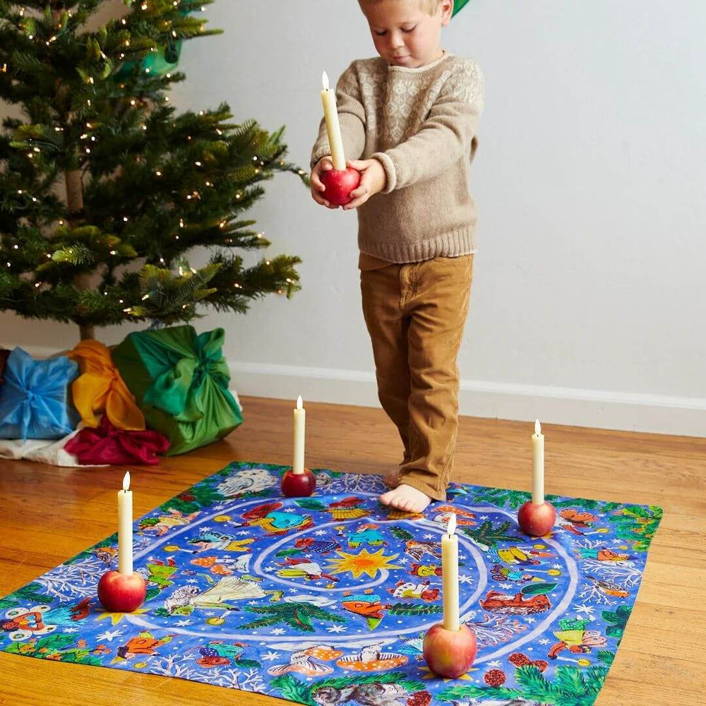 A child holding a candle walks on the Phoebe Wahl illustrated advent spiral playsilk by Sarah's Silks.