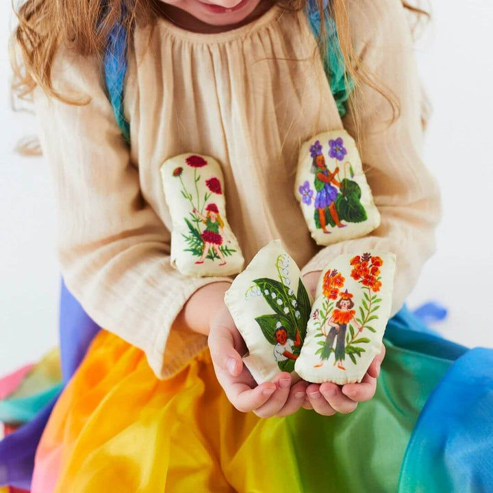 A child in a silk rainbow skirt holding all four of the Sarah's Silks Stuffed Flower Fairy figures, featuring Phoebe Wahl’s whimsical illustrations on soft silk-covered pillows.