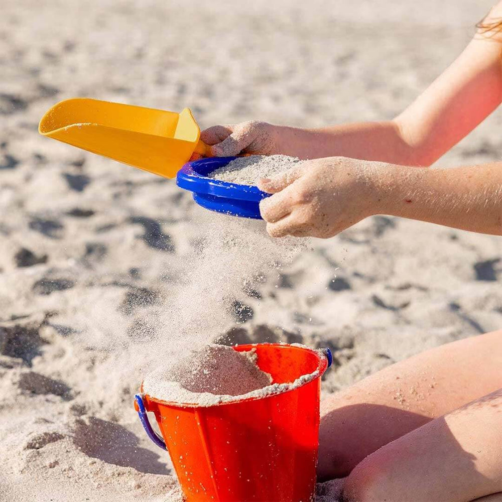 Child sifting sand through blue sand sieve into a red pail, while holding a yellow sand scoop on the beach