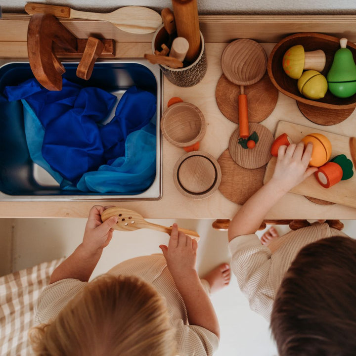 Bella Luna Toys- Children playing with wooden play food on wooden play kitchen counter- Play kitchen counter includes sink with wooden faucet, and counter full of wooden kitchen tools