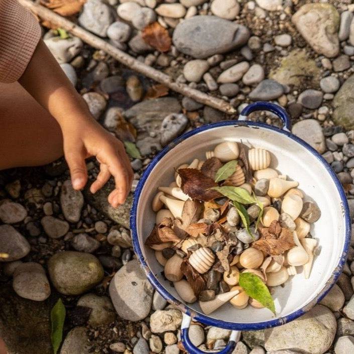 Child playing on a stony beach with a pot of Grapat Mandala Natural Wooden Shapes, and some leaves. Maybe a "stone soup."