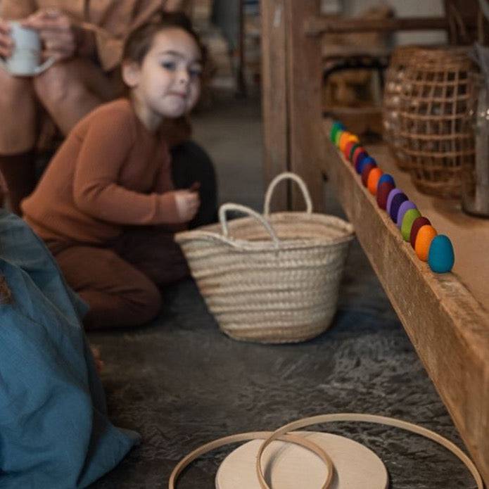 Adult and child crouched down in background, looking towards Grapat Mandala Wooden Rainbow Eggs lined up along shelf nearby